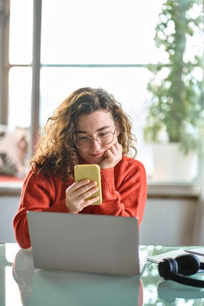 Eine junge Frau in rotem Pullover schaut lächelnd auf ihr Smartphone am Tisch sitzend.